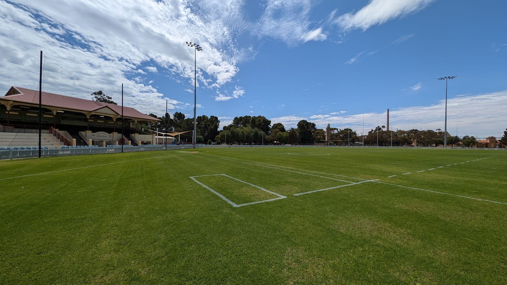 A large, well-maintained sports field with green grass and white line markings. On the left, there is a grandstand with a red-tiled roof and several rows of seating. Tall floodlights are positioned around the field. In the background, there are trees and some buildings, including a tall chimney-like structure. The sky is partly cloudy.
