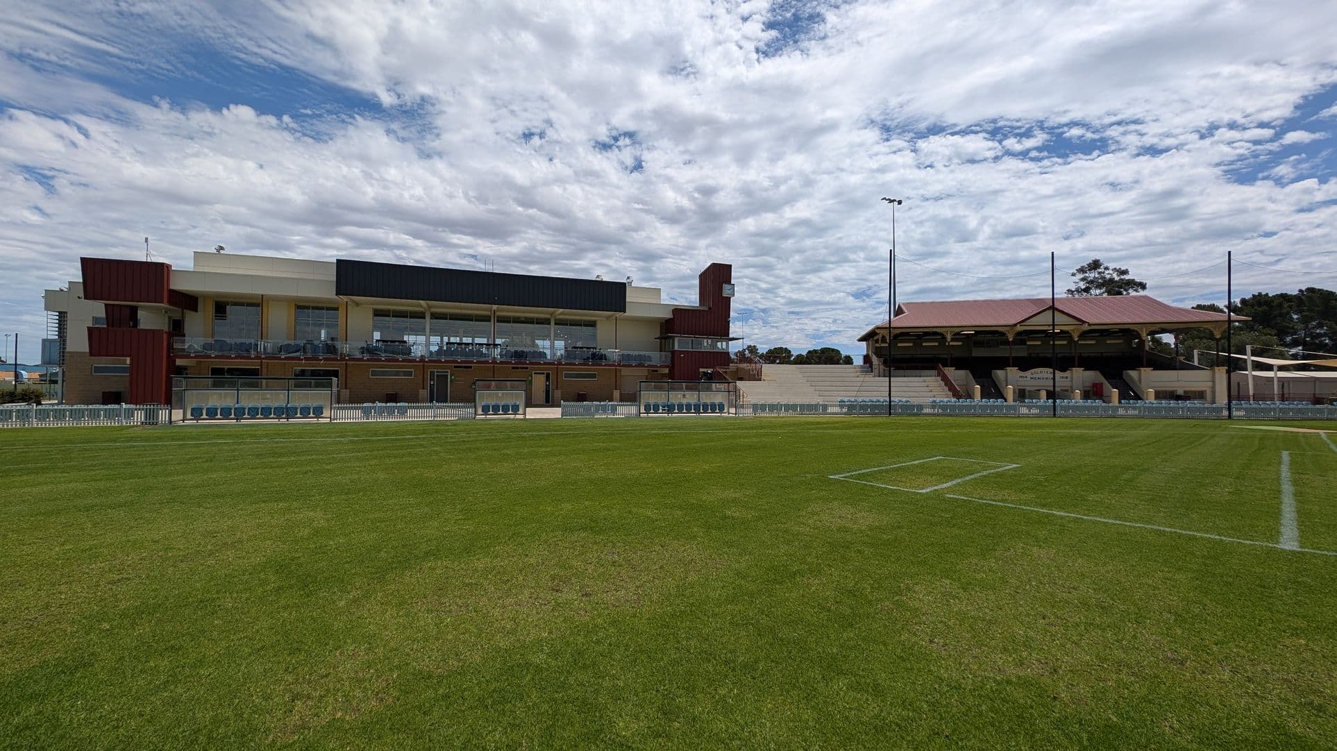 A sports field with well-maintained green grass in the foreground. On the left, there is a modern building with large glass windows and a mix of brown and cream colours. In the centre, there are seating areas with blue chairs behind a white fence. On the right, a traditional grandstand with a red roof and wooden supports. Tall floodlights are visible, and trees are in the background.