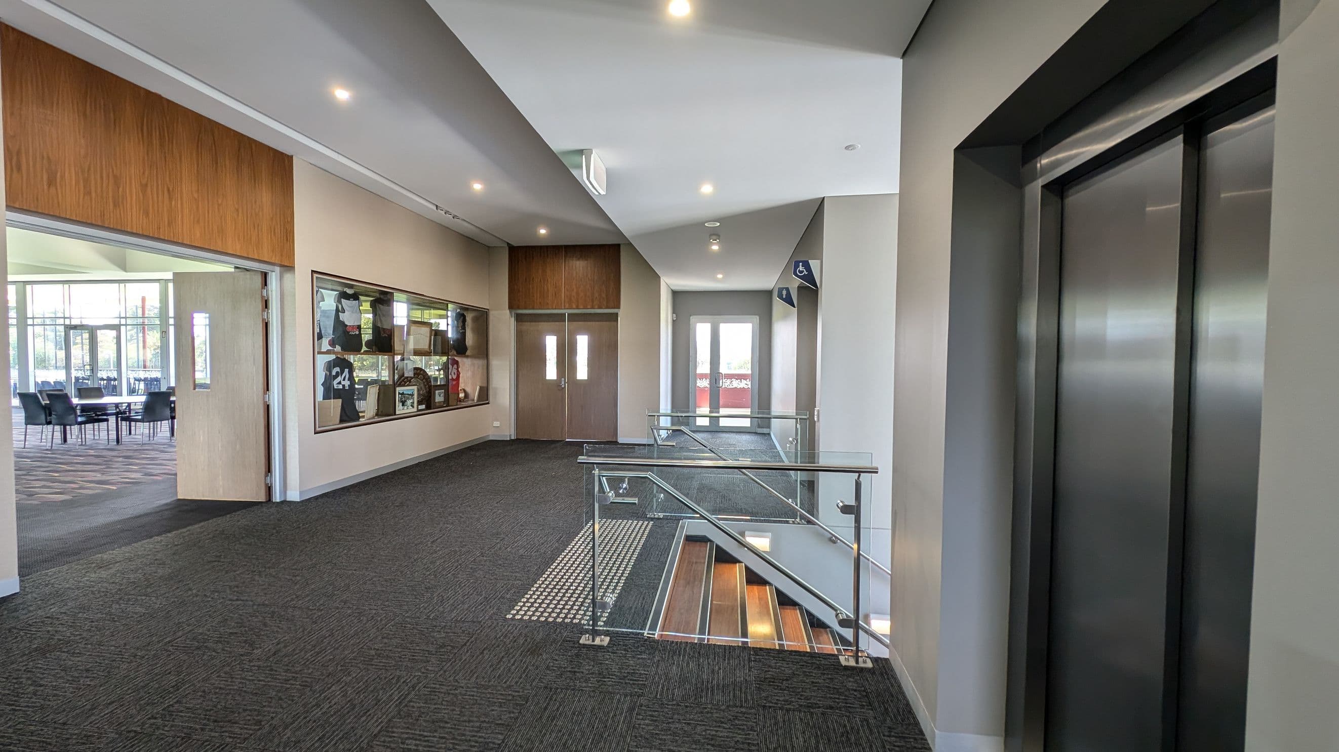 A modern indoor hallway with grey carpeted flooring. On the right, there is a closed lift with metallic doors. In the centre, a glass railing leads down a staircase. On the left, a display case with sports memorabilia, including jerseys, is mounted on the wall. Further left, an open doorway reveals a room with tables and chairs. The ceiling is white with recessed lighting, and there are double doors at the end of the hallway.