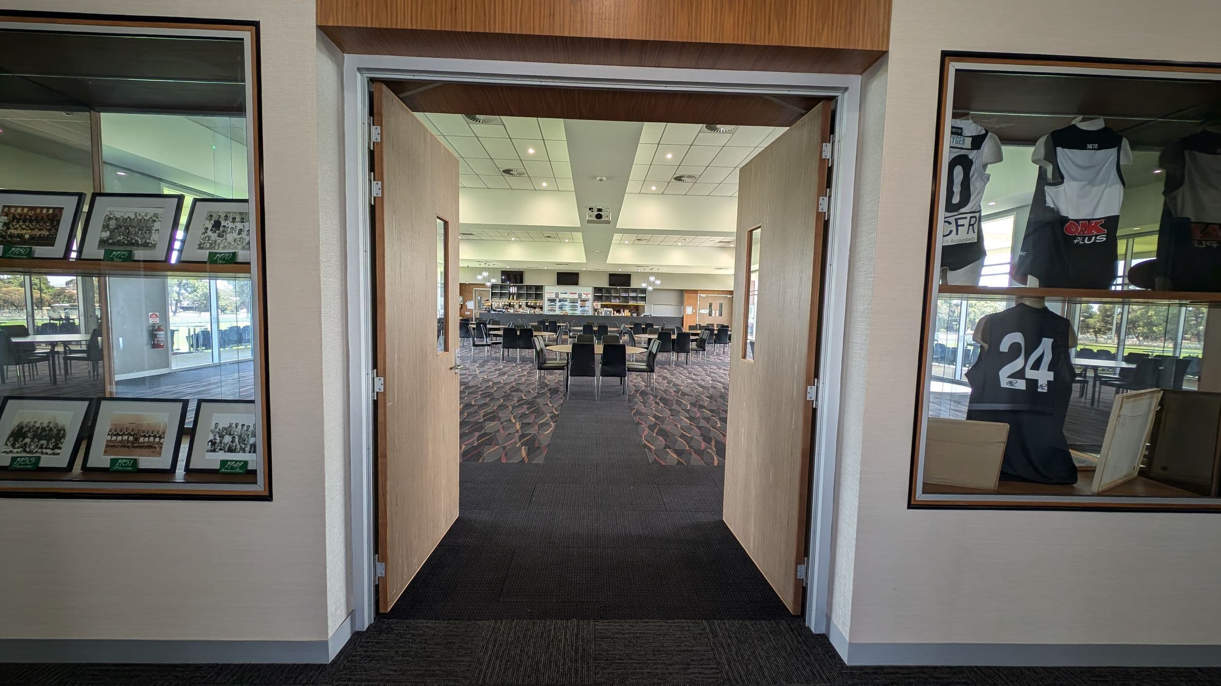 A hallway with open double doors leading to a spacious room. On the left, a display case contains framed photographs. On the right, another display case showcases sports jerseys, including one with the number 24. The room beyond has a patterned carpet, tables, and chairs, with a bar or counter area at the far end. The ceiling is fitted with lights and a projector. Large windows on the left provide natural light.