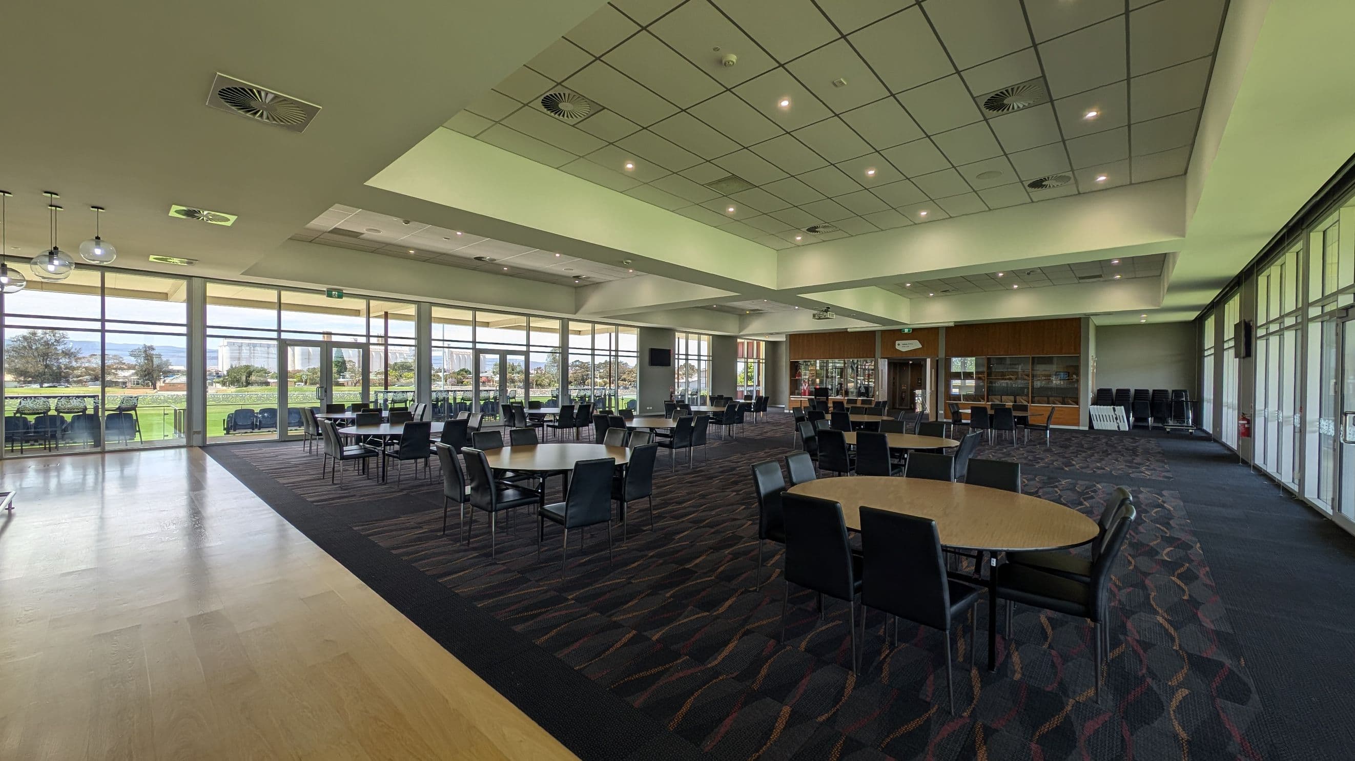 A spacious room with large windows on the right, offering a view of a green outdoor area. The room has a mix of wooden and carpeted flooring. Several round tables with black chairs are arranged throughout the space. The ceiling is fitted with recessed lighting and air vents. At the far end, there is a wooden wall with glass display cabinets and a doorway. The left side features hanging spherical lights.