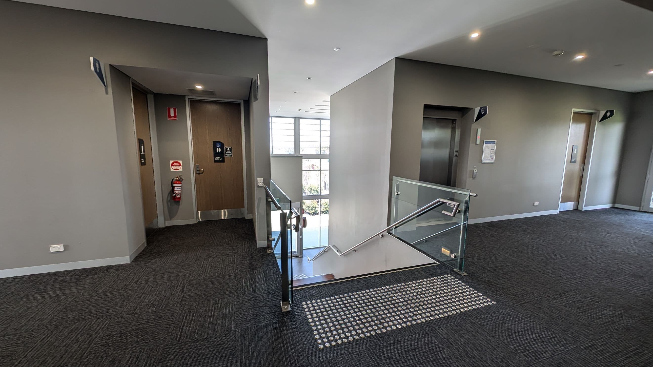 A modern indoor space with two lifts on either side of a central staircase. The left lift is next to a wooden door with a fire extinguisher mounted on the wall. The right lift is adjacent to a notice board. The staircase has glass railings and leads downwards, with a large window in the background allowing natural light. The floor is carpeted in dark grey, and the ceiling is fitted with recessed lights.