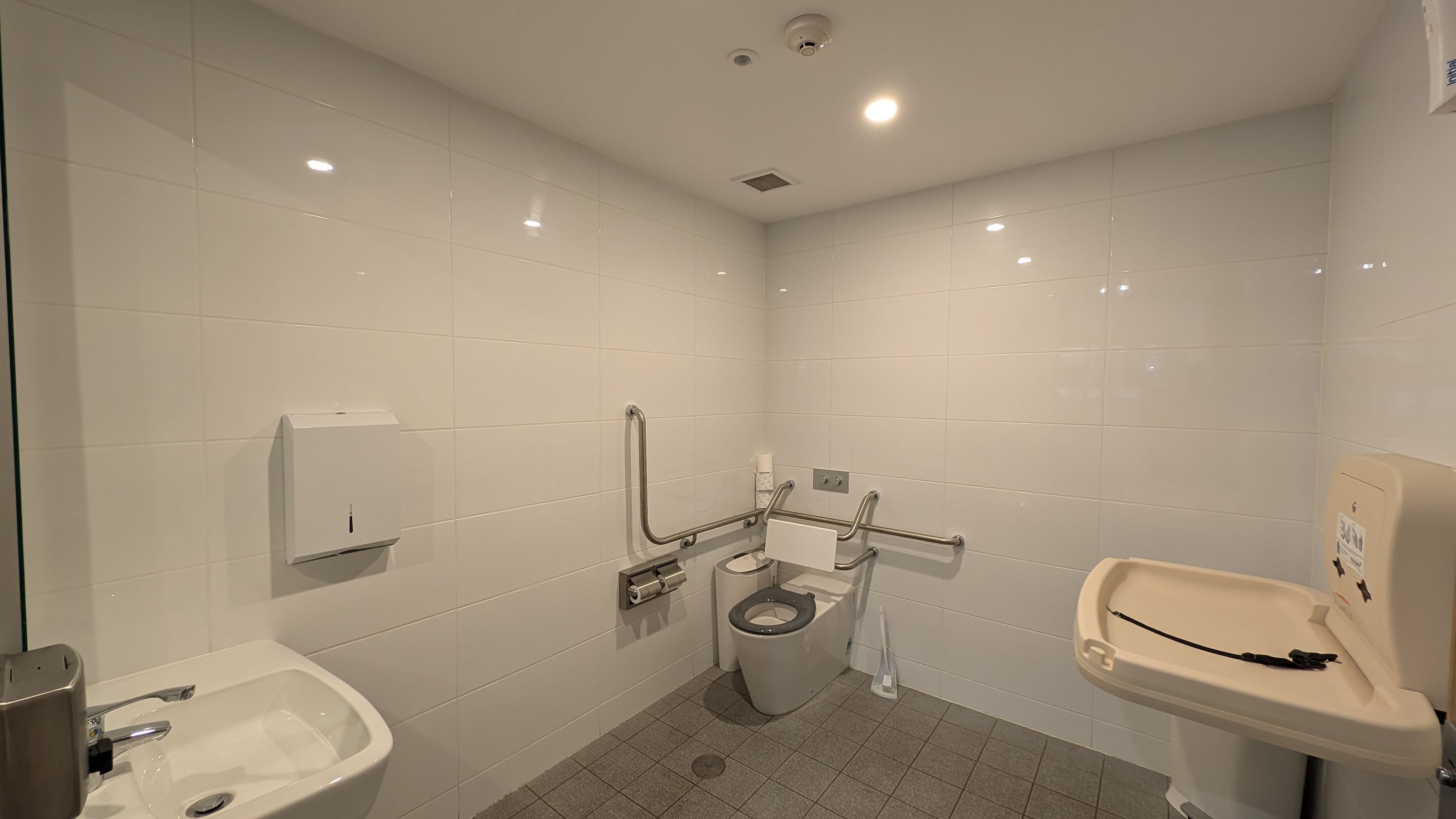 A public accessible toilet with white tiled walls and a grey tiled floor. In the centre, there is a toilet with support rails on both sides. To the left, a small sink with a soap dispenser and a paper towel dispenser above. On the right, a baby changing station is mounted on the wall. The ceiling is white with recessed lighting and a smoke detector.