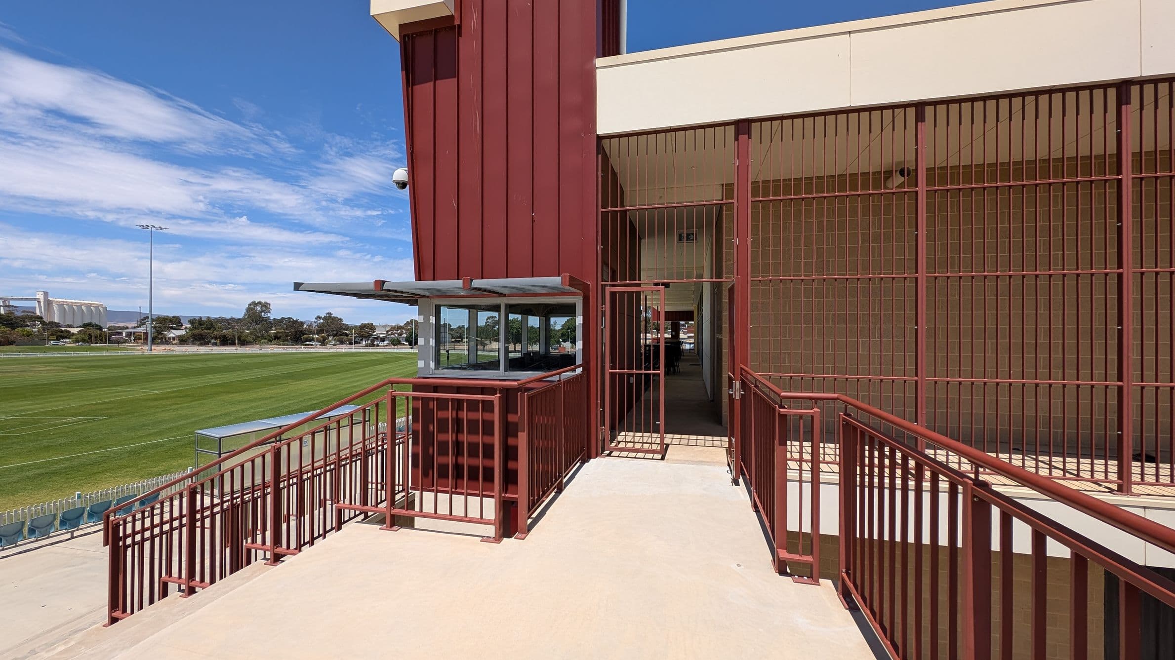 A sports field with neatly mowed grass is on the left, featuring a few rows of blue seating and a small shelter. On the right, a building with red metal railings and a gated entrance is visible. The building has a beige upper section and a red vertical panel. A security camera is mounted on the corner. In the background, there are trees and industrial structures. The ground is paved with light-coloured concrete.