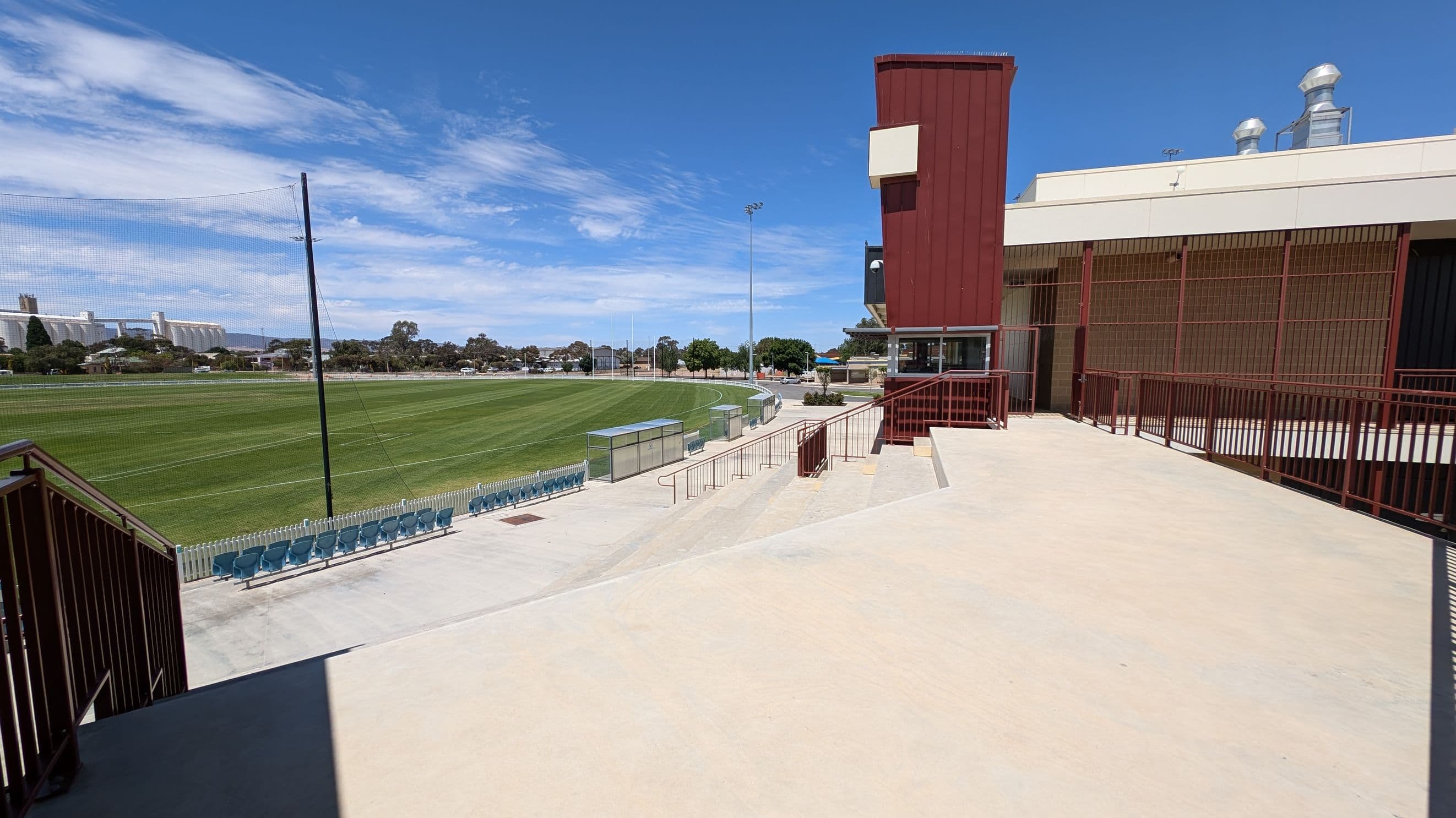 A sports field with a well-maintained green lawn is on the left, surrounded by a white picket fence and rows of blue seating. In the centre, a pathway leads towards the field, with a few trees and buildings visible in the distance. On the right, a red and beige building with metal railings and a ramp is present. The building has a flat roof with ventilation ducts. The foreground features a light-coloured concrete surface.