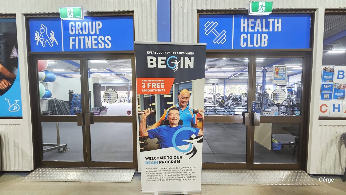 Group Fitness and Health Club at Portland Leisure and Aquatic Centre. Both facilities are distinguishable by the rectangular metals with bumps and a large blue sign with two little green EXIT signs on top of the manually operated glass double doors with chocolate metal frames. The cream wall has corrugated linings. A tall, vertical banner with a blue background and white text stands out between the doors.