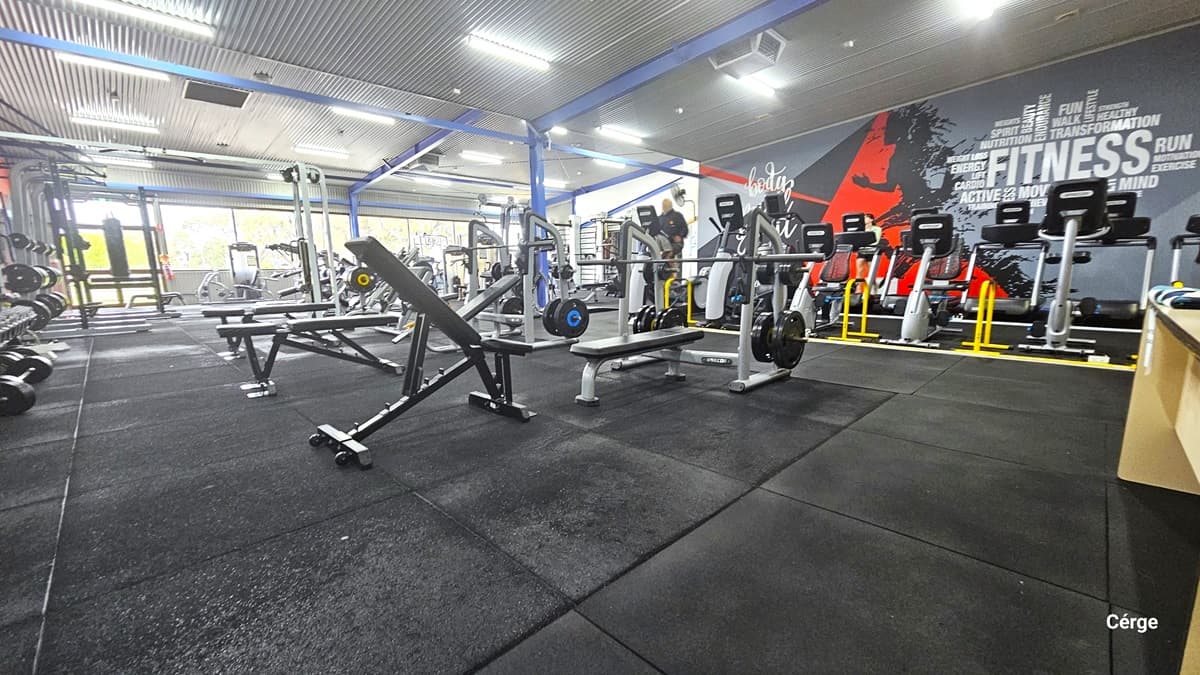 Inside the Health Club. Black rubber mats cover the floor, hosting a variety of exercise equipment in black, silver, red, and yellow accents. Weight racks, benches, and cardio machines are visible. A striking feature is a large grey mural on the right wall, painted in red and black, with the words “FITNESS” and “RUN” prominently displayed. The room has blue metal beams and a grey corrugated ceiling and is well-lit by fluorescent lights.