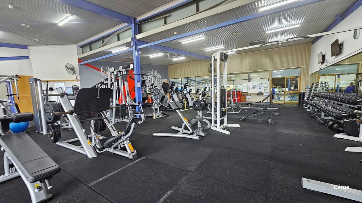 Leg bench presses, stationary bikes, dumbbells, blue exercise balls, and other exercise equipment inside the Health Club. The weight racks with dumbbells are placed near the large wall mirror to the right white wall with a clock and mounted flat-screen TVs. The entrance glass windows and doors have a beige corrugated wall. Mounted on the white wall after the grey mural is a wall fan. The room has blue metal beams and a grey corrugated ceiling and is well-lit by fluorescent lights.