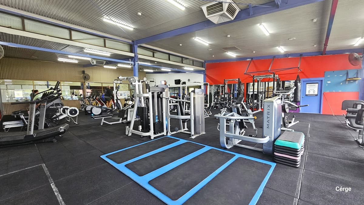 Blue rectangular rubber mat with smaller black mats. The high-ceilinged space is well-lit, revealing a variety of exercise equipment on gray rubber mats. The equipment, mostly black and silver, includes treadmills, weight machines, and free weights. A blue metal beam, door, and red wall are evident on the back wall. Further to the left are the stationary bicycles and treadmills.