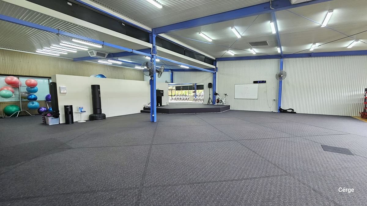 Large, well-lit Group Fitness Centre. The floor is covered with gray mats, and the gym is adorned with a variety of exercise equipment. On the left, exercise balls and other equipment are neatly stacked against the wall. On the right, a whiteboard and a stationary bicycle are visible. At the center is a short black stage with a small speaker placed on a tripod. Wall fans are placed on the blue metal beams.