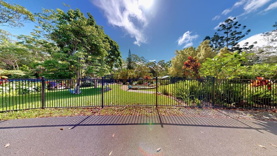 A black metal fence divides the aquatic area from the road. Inside, pools are visible. Plastic chairs are under tree shades on the left, while various trees, ferns, and ornamental plants decorate the landscape on the right.