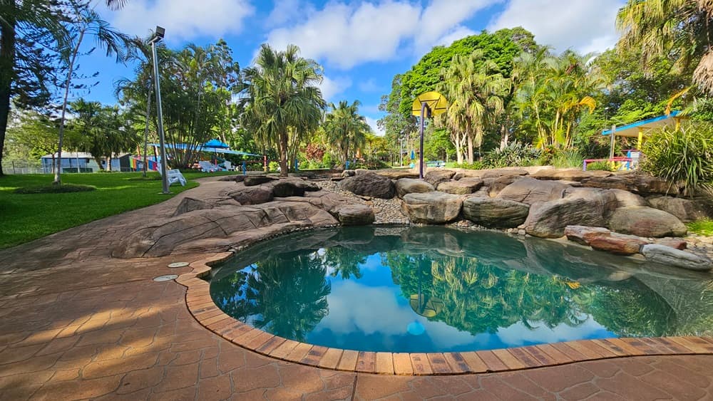 A circular pool is bordered by large rocks and pebbles. Plastic lounge chairs are placed on the grass area to the left, alongside palm trees. Trees and grass encircle the area. A basketball board is on the pool deck. On the right, there's a colourful shade structure. Trees surround the area.