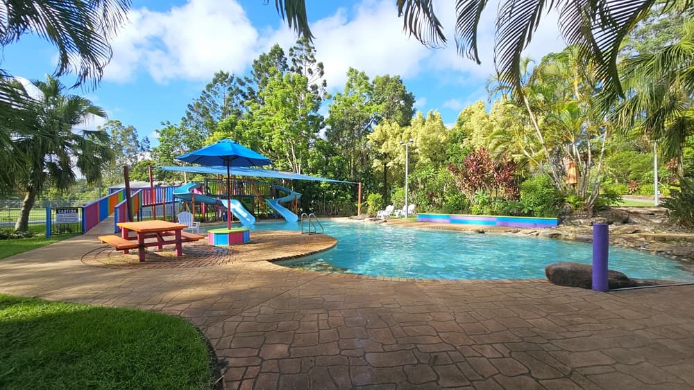 A children's pool with handrails and steps. Blue slides with multicolored ramps on the left, featuring safety reminders. A picnic table and umbrella shade on the pool deck. A large rock and a purple bollard are visible. Surrounding the area are various trees and grass. Plastic lounge chairs are positioned on the other side of the pool.