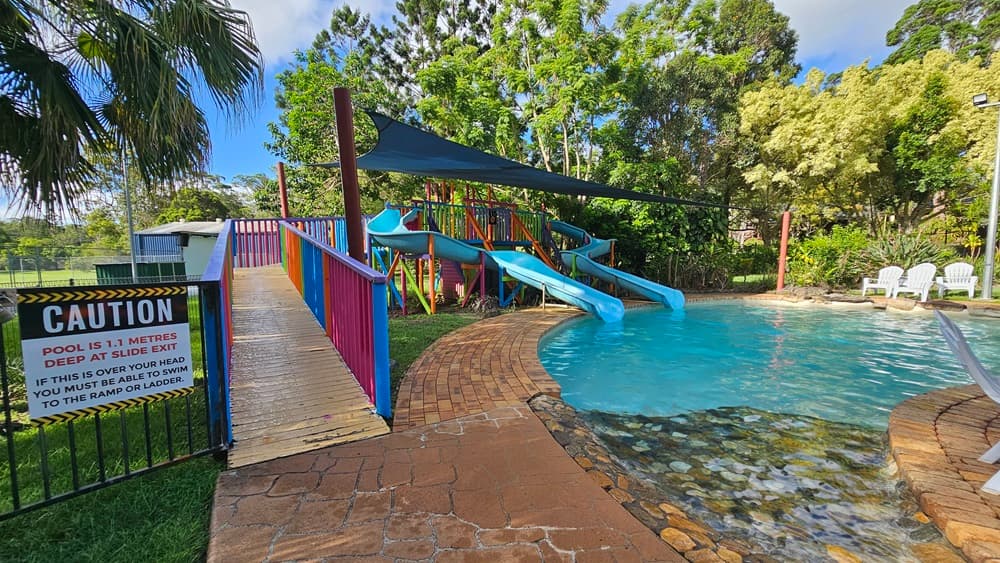 A children's pool with handrails and steps. Blue slides with multicolored ramps on the left, featuring safety reminders. A black shade cloth hangs overhead. Surrounding the area are various trees and grass. Plastic lounge chairs are positioned on the other side of the pool.
