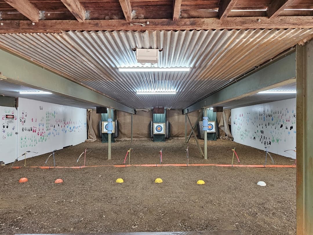 On the left and right, white walls are covered with various colourful handprints and drawings. In the middle, there are three archery targets mounted on stands, each with a blue and white bullseye. The background features a beige netting behind the targets. The foreground has a dirt floor with a row of yellow, white, and orange cones. The ceiling is corrugated metal with exposed wooden beams.