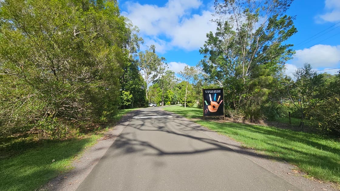 A straight bitumen road. Alongside the road, there is a large sign situated on the grassy area to the right. The road is flanked on both sides by various trees with robust branches and grass.