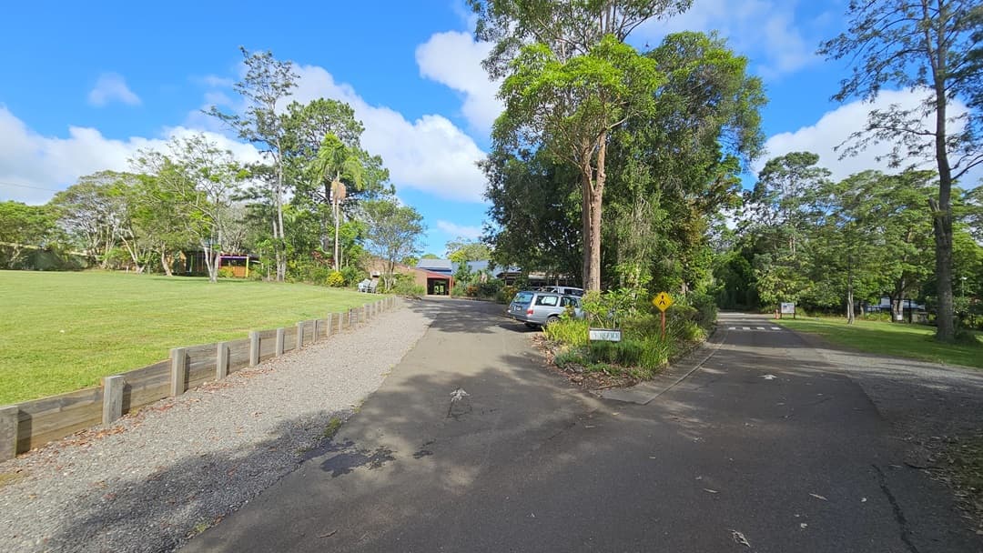 The road splits: To the left, it leads to a car park and a building with a brown brick facade, alongside a field of manicured grass bordered by a low concrete fence. The right-hand road leads to an open area surrounded by trees and grass. In the middle aisle, there is a sign, set against a backdrop of tall trees and grass.