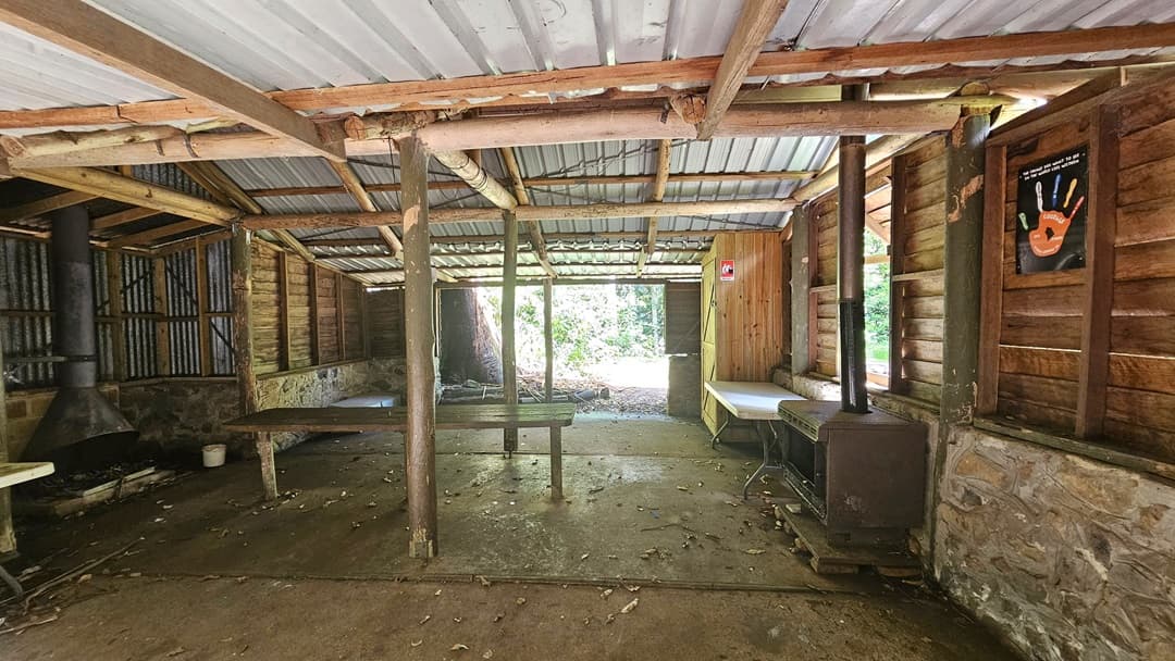 A hut with a half-concrete, half-timber structure. The corrugated roof and wooden beams make up the ceiling. A poster hangs on the right timber wall. Tables, bench chairs, and a cabinet are arranged against the wall. A fireplace and chimney are on the left. The uneven floor is hard concrete with scattered dry foliage.