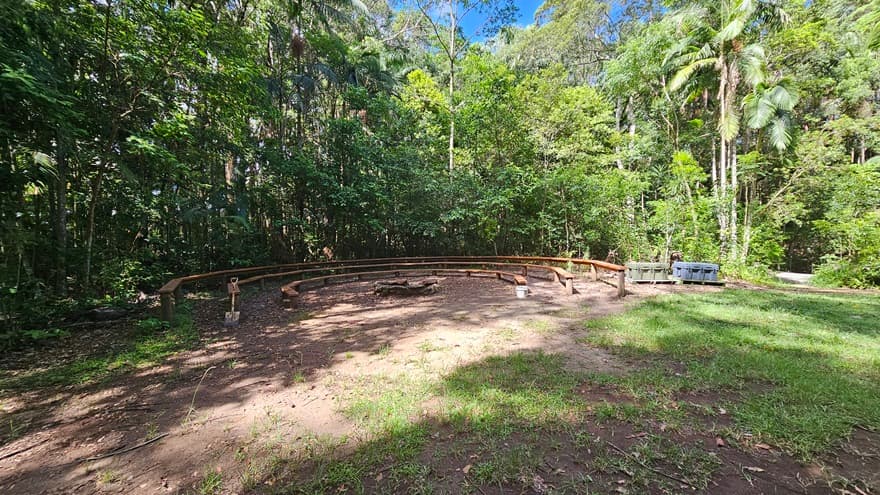 In an open unpaved area partially covered with grass, wooden bench chairs form a U-shape on the left. Tall trees, bushes, native plants, and grass, including palm trees, border the back.