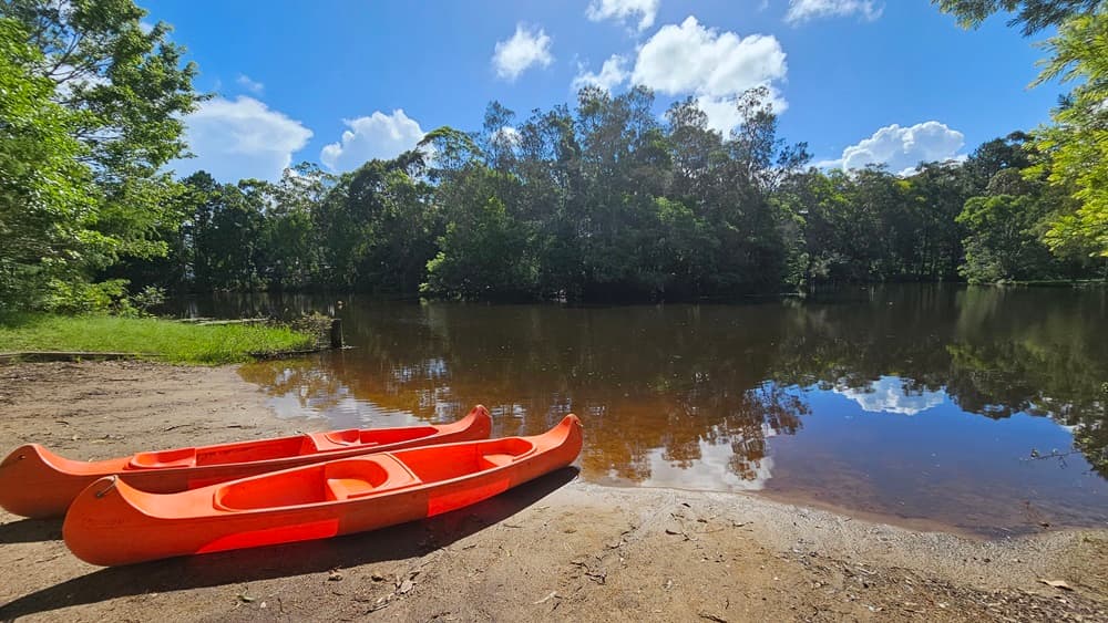 Two orange watercraft rest near the lake, their tips touching the water. Surrounding the lake are various trees and native grasses.