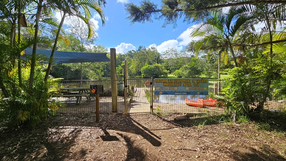 Bench chairs are neatly arranged beneath a shade cloth secured by a metal fence displaying posters and facility reminders. A wooden platform extends from the shade to the lake. Orange watercraft rest by the lake on the right, bordered by palm trees in the front and various trees surrounding the area. The ground is unpaved.