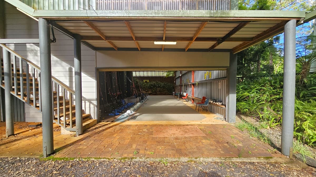 To the left is a staircase with metal handrails. Under the shade of the tower are climbing gear such as ropes and helmets positioned on the floor in front of the black climbing wall. Plastic chairs are lined near the corrugated wall, along with headgear. Landscape plants fill the end. The area on the right is filled with native grass, ferns, and trees. The floor has concrete and bricks.