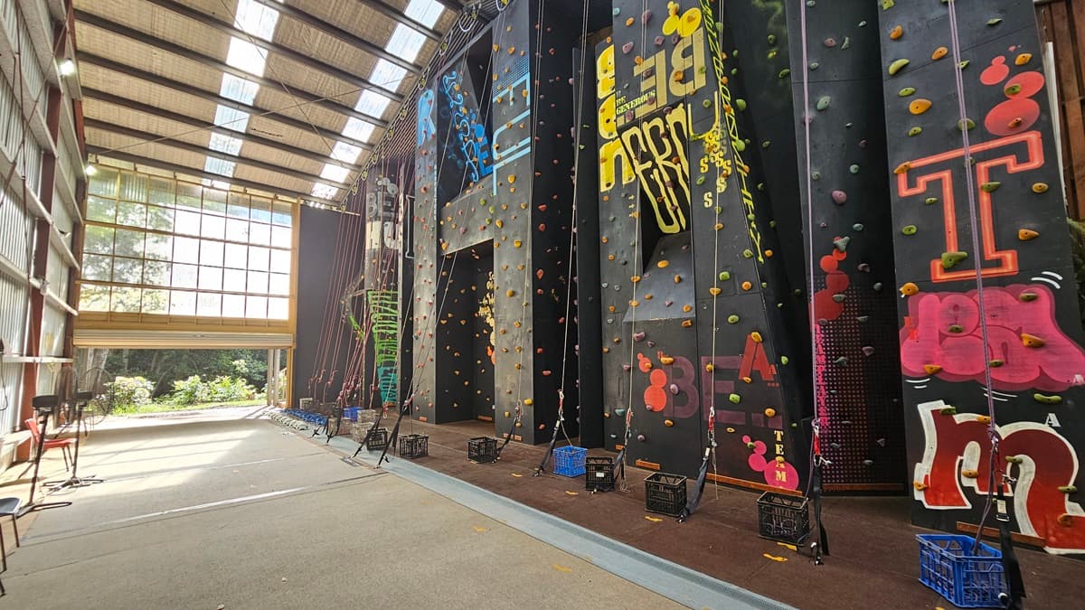 To the right, a black climbing wall with patterns and multi-colored steps is equipped with climbing gear like ropes, headgear, and gloves. The ceiling and upper wall display large metal beams with silver insulation. The left wall has corrugated metal sheets. Plastic chairs are arranged, along with a fan. The industrial door is rolled up at the top.