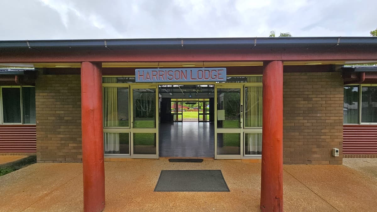A road with white markings on the left, flanked by trees and bollards. On the right, a low-rise building with a brown brick facade and pitched, extended roofing. Well-manicured green grass lines its left side. Trees and shrubs surround the area.