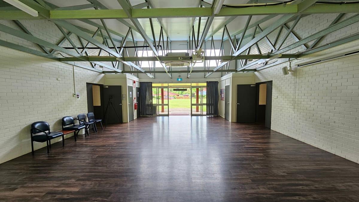 A spacious open area with a brown timber floor. Black plastic chairs arranged against the left wall. White brick wall and white ceiling with exposed metal beams. Black outward-opening doors on both sides lead to corridors. Further back, green grass is visible.