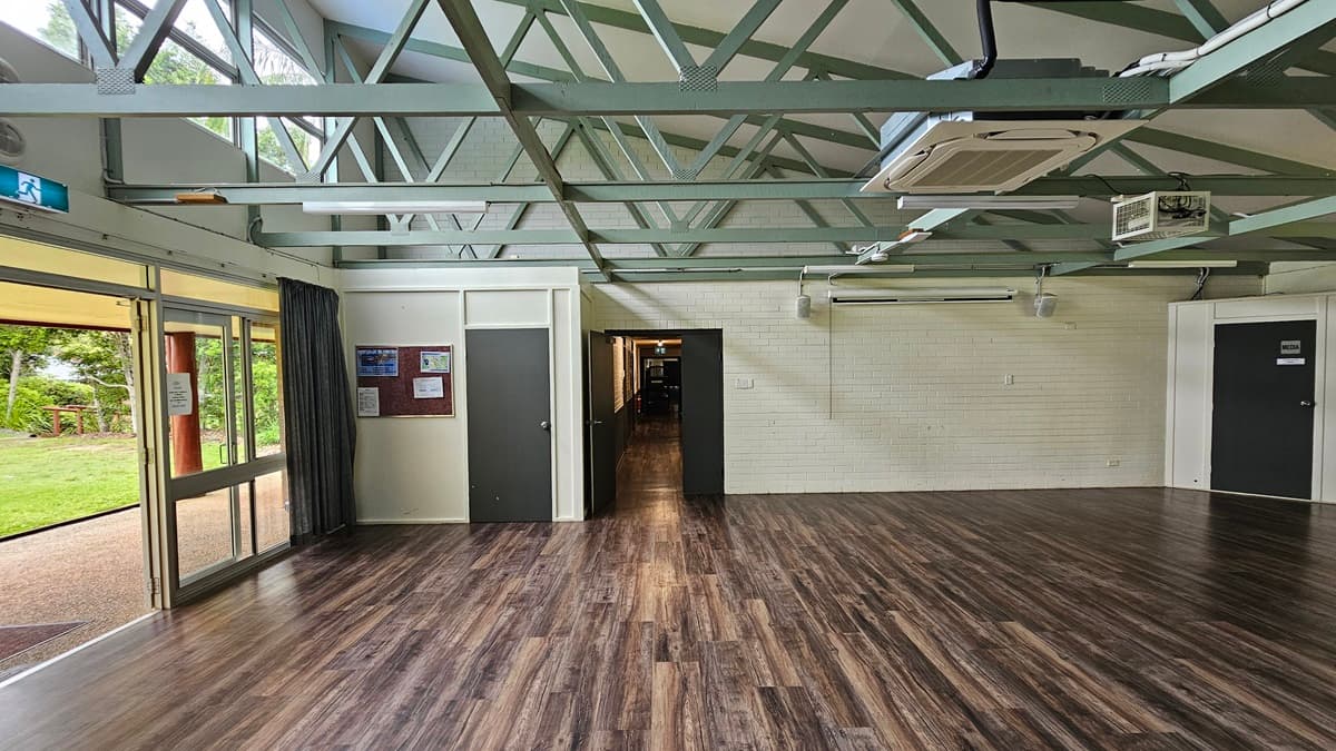 A spacious open area with a brown timber floor. On the left. a white brick wall holds a brown board with posters. The white ceiling features exposed metal beams, fluorescent lights, and air conditioning. A black door on the left leads to a corridor, and another black door is on the right. To the left, the open glass door exposes green grass outside.