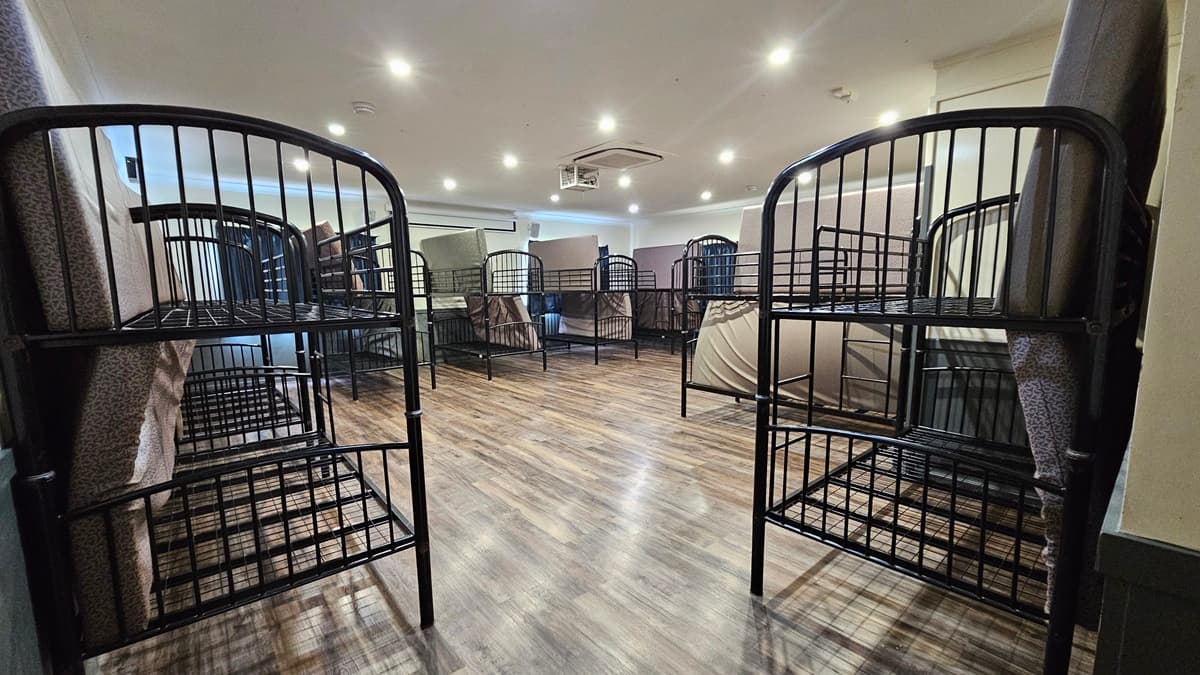 A room with a brown timber floor featuring metal bunk beds strategically positioned against the walls. Each bed has mattresses laid at the sides. The white ceiling has LED lights and air conditioning, and the wall is white.