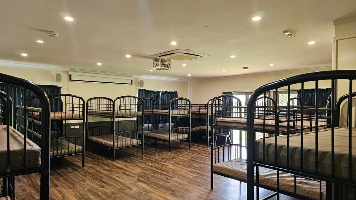 A room with a brown timber floor featuring metal bunk beds strategically positioned against the walls, each equipped with mattresses. The white ceiling has LED lights, air conditioning, and a projector machine. The wall is white with a projector board. Windows are covered with dark blue curtains.