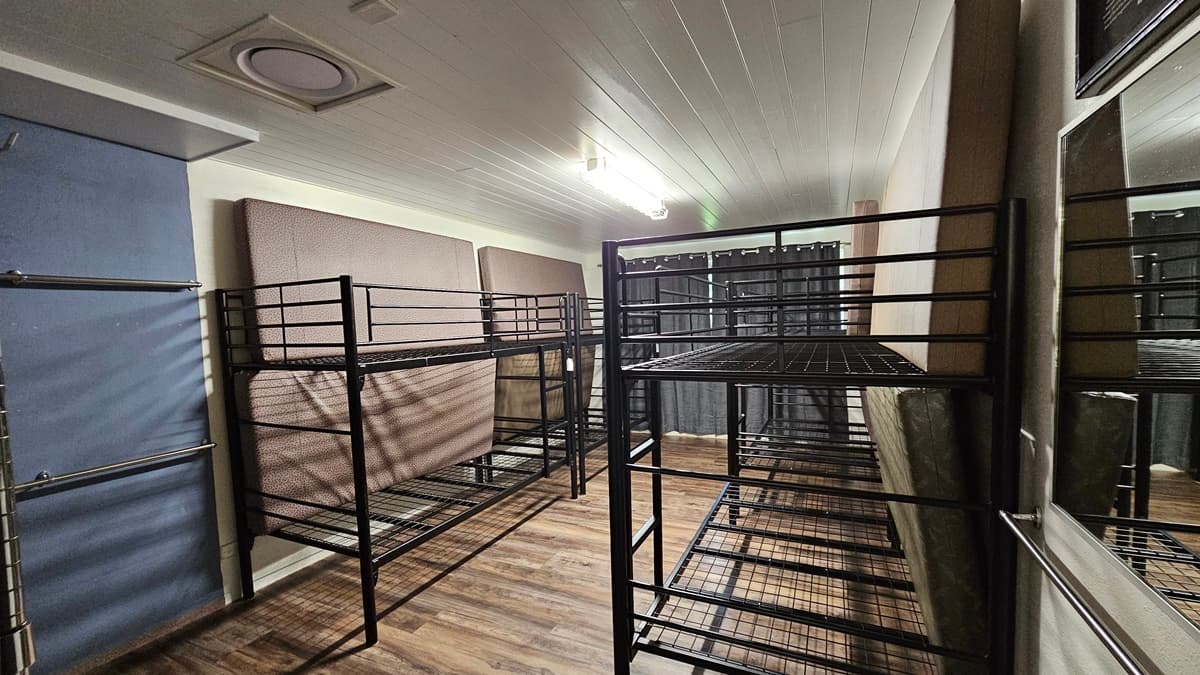 A room with a brown timber floor featuring black metal bunk beds strategically positioned against the walls, each equipped with mattresses laid on the sides. The white ceiling has LED lights. The wall is all white.