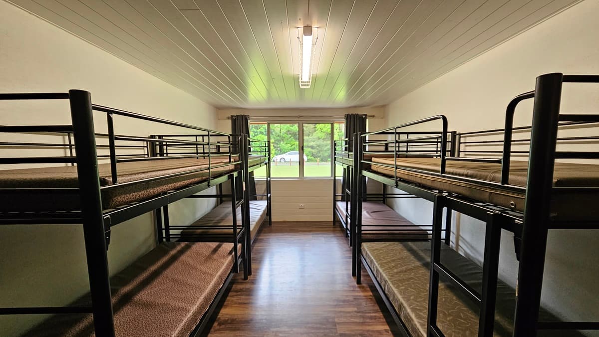 A room with a brown timber floor featuring black metal bunk beds strategically positioned against the walls, each equipped with mattresses. The white ceiling has a fluorescent light. In the middle is a glass window with dark curtains rolled to the sides, offering a view of the green grass outside and a white car.