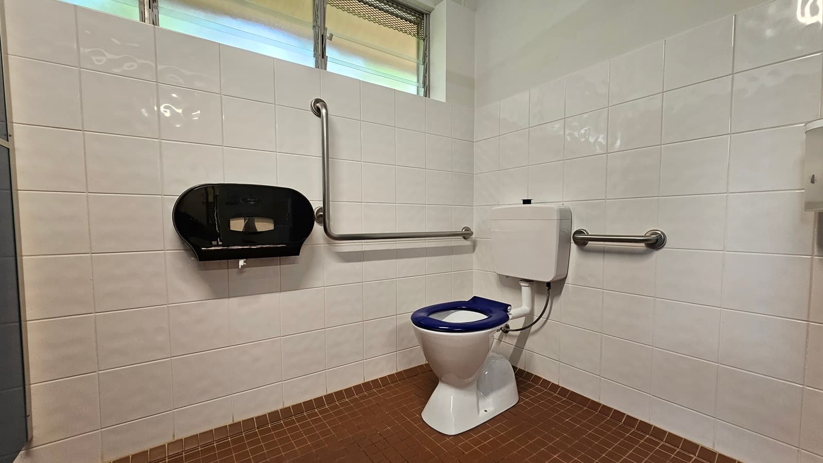 In the corner, a toilet bowl with handrails. A tissue dispenser is on the left wall. Glass jalousie windows are on the upper left, and a liquid soap dispenser is on the upper right wall. White tiles cover the walls, while the floor is tiled brown.