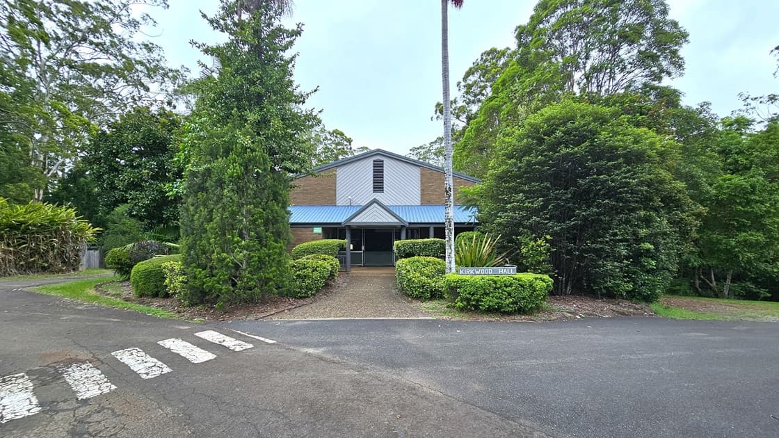 In the middle is a low-rise facility with a pitched roof, and a brown brick facade with white and blue trims. Trimmed shrubs border the front and sides. A sign is placed next to a tall wooden post on the right. Various trees surround the area and border the road with white markings for pedestrians.
