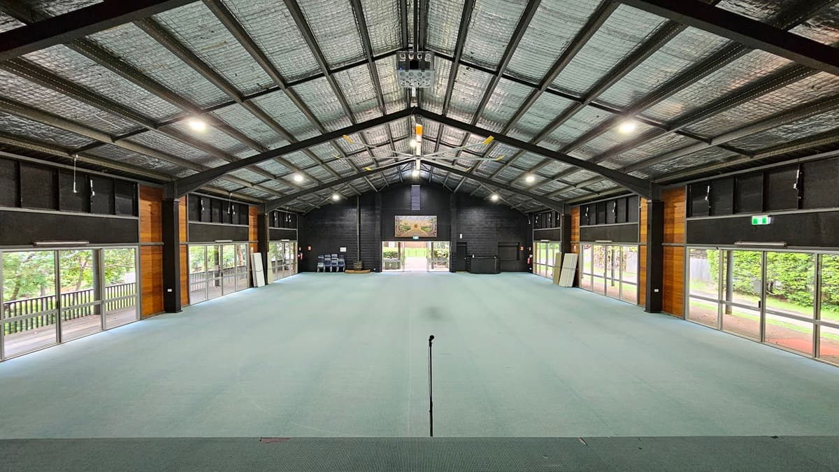 A spacious open area with a pitched industrial-style ceiling, exposed metal beams, silver insulation, a large ceiling fan, and a projector machine. Both sides have large glass windows and doors offering a view of trees outside. The walls feature black and brown timber, and the floor is covered with grey tiles. Plastic chairs are piled in the left corner, and a large flatscreen TV is mounted on the middle wall parallel to the entrance.