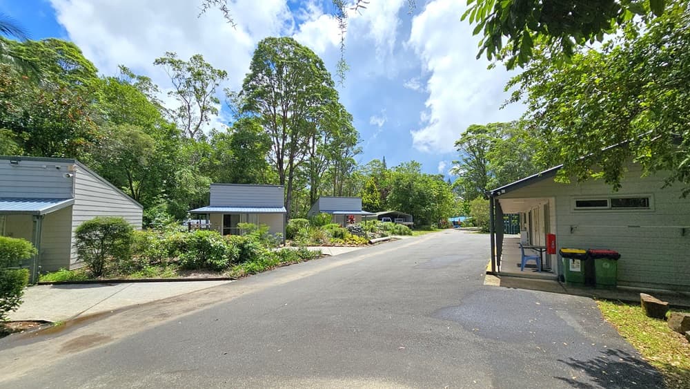 In the middle is a concrete road. Low-rise buildings with concrete walls and corrugated roofing are positioned on both sides. The longer building on the right displays a fire extinguisher on the wall, along with a table, chair, and two rubbish bins. Trees and grass surround the area.