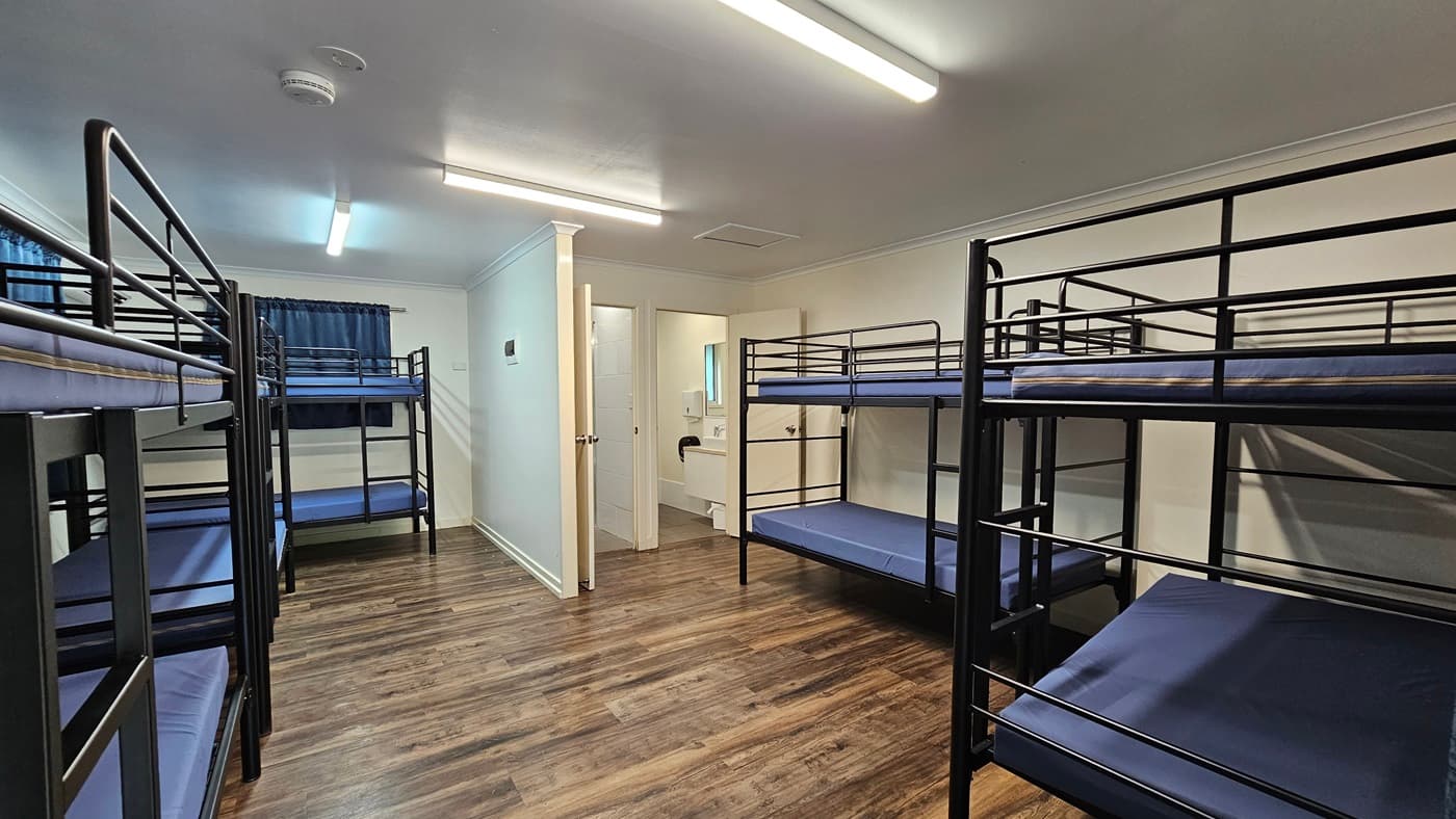 A room with metal bunk beds, each with mattresses. Large windows with grey curtains rolled to the sides. A mirror and signage are mounted on the middle wall. White walls and ceiling with fluorescent lights and air conditioning. Brown timber floor.