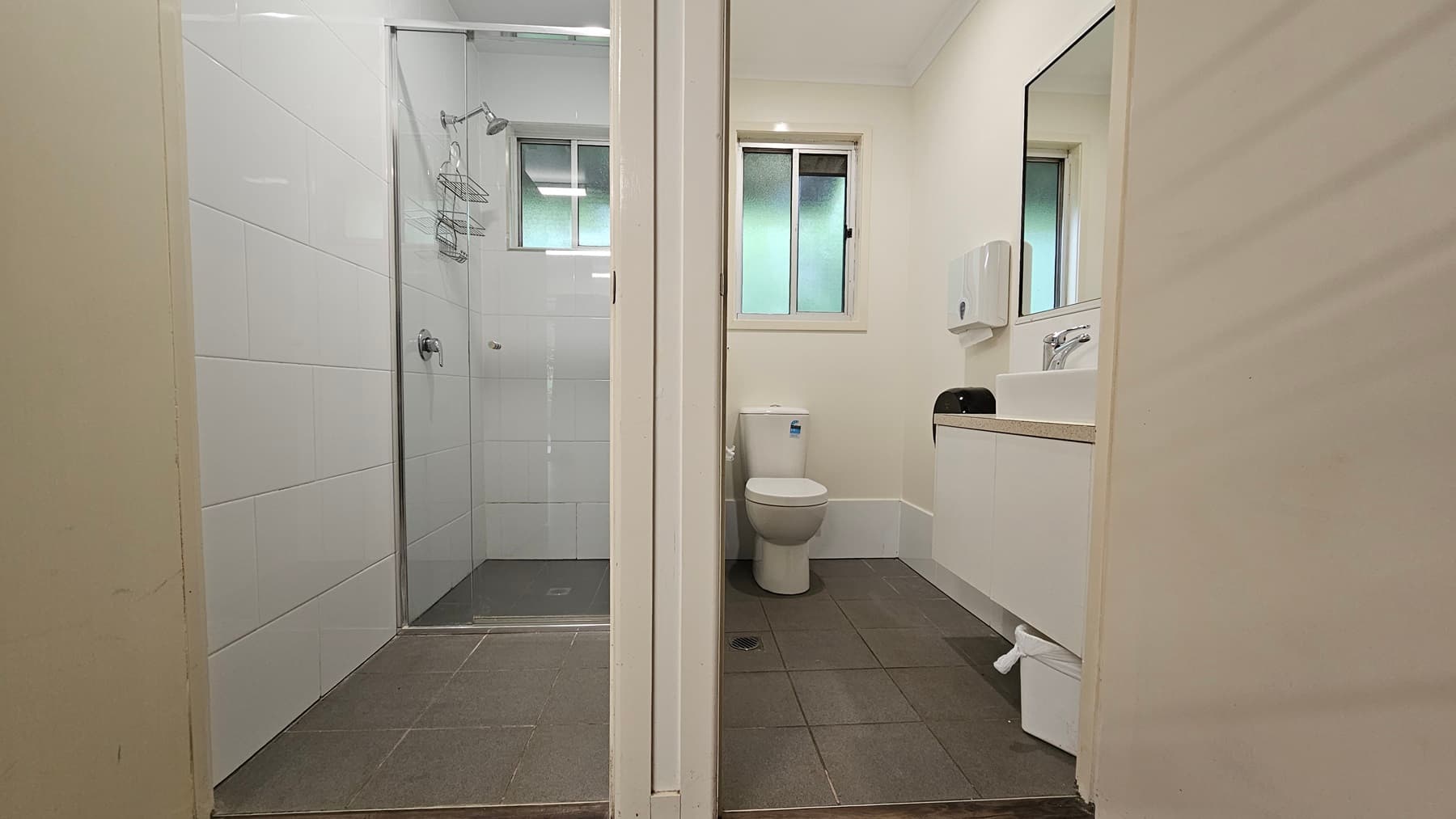 Two separate cubicles. To the right, a toilet bowl is positioned in the corner with a glass window above. On the right wall, a mirror, tissue dispenser, and hand basin are mounted. A rubbish bin is placed underneath. To the left is a shower area enclosed with glass. Both cubicles feature white walls, grey floors, and small windows.