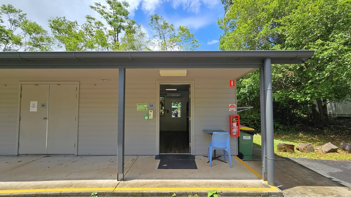 A low-rise building with extended roofing and white brick walls with multiple doors. The open door on the right reveals timber floors and a mirror inside. The outer wall features a fire extinguisher and signage. A table and plastic chair are placed to the right of the door. Trees and grass surround the area, and rubbish bins visible on the right.