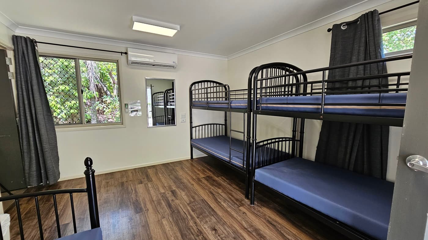 A room with metal bunk beds, each with mattresses. Large windows with grey curtains rolled to the sides. A mirror and signage are mounted on the middle wall. White walls and ceiling with fluorescent lights and air conditioning. Brown timber floor.