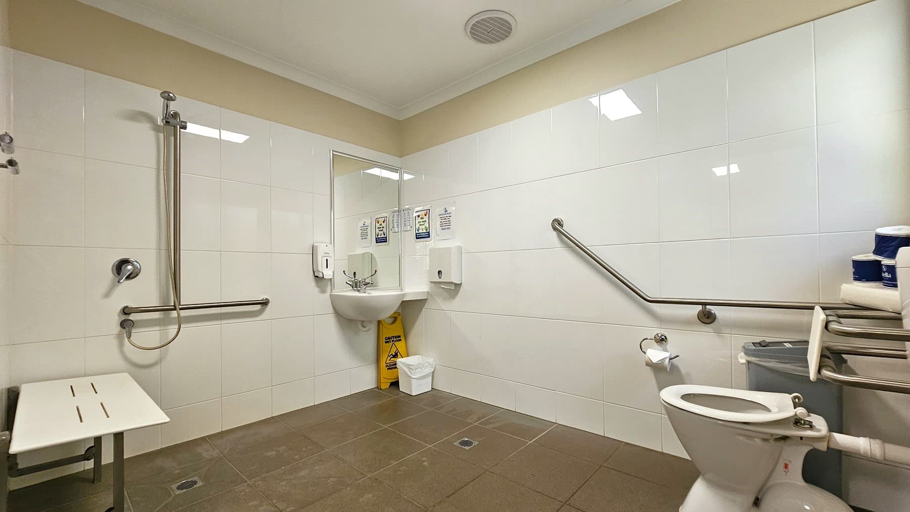 A toilet bowl equipped with handrails on the right. Tissue rolls and a rubbish bin are positioned to the right. In the middle corner is a hand basin. There is a tissue dispenser, liquid soap dispenser, and stickers. A rubbish bin and a yellow warning sign are underneath. Adjacent is a shower with handrails, and next to it is a foldable bench chair mounted to the left wall. The ceiling is white with a cream accent, and the walls are tiled with white tiles, while the floor has brown tiles.