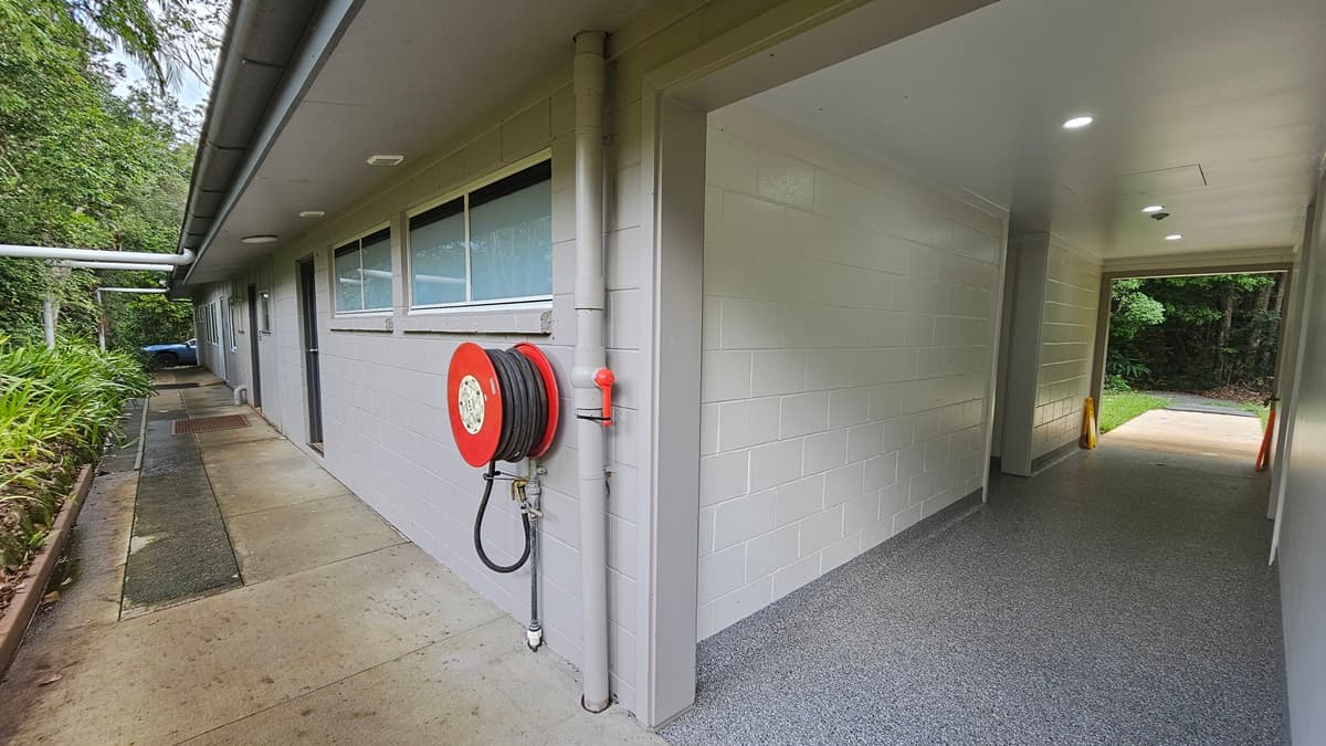 A low-rise building with a white brick facade, multiple doors, and glass windows. A fire hose reel is mounted near the corner. Landscaped ornamental plants and trees border the left side. The lobby on the right has grey tiles and leads to the other side of the building. Safety warning signs are placed near the wall.