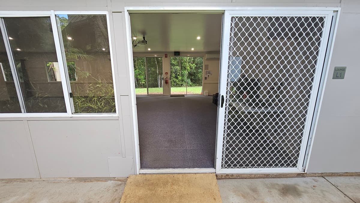 A glass door with a security screen and a yellow mat. Adjacent are glass windows. Inside is a spacious open space with grey flooring and a white ceiling. Signs, along with a fire extinguisher, are mounted on the middle wall, next to another glass door that leads to the green field outside.
