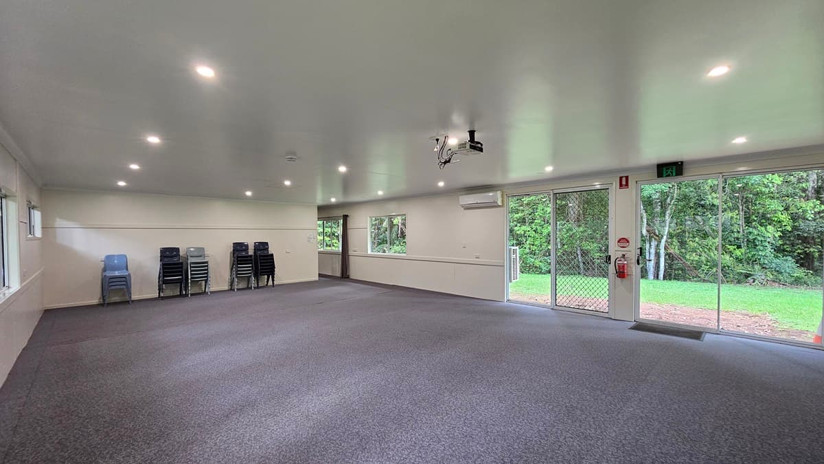 A spacious open space with grey flooring and a white ceiling. Signs and a fire extinguisher are mounted on the middle wall, next to a glass door leading to the green field outside. A projector machine is mounted to the ceiling along with LED lights. Plastic chairs are piled against the middle wall. Trees are visible on the right.