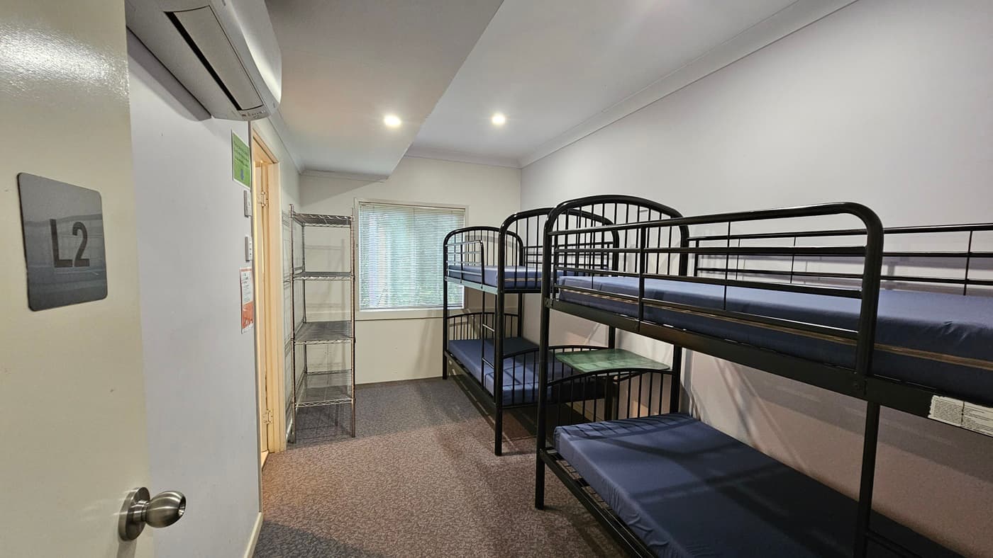A room with metal bunk beds, each with mattresses, positioned against the wall. On the left is a door, adjacent to a 5-tier movable stainless display rack against the wall. In the middle wall is a glass window with blinds. The walls are white with poster reminders. An air conditioner is mounted to the upper left wall. The ceiling is white with fluorescent lights, and the floor is brown timber.