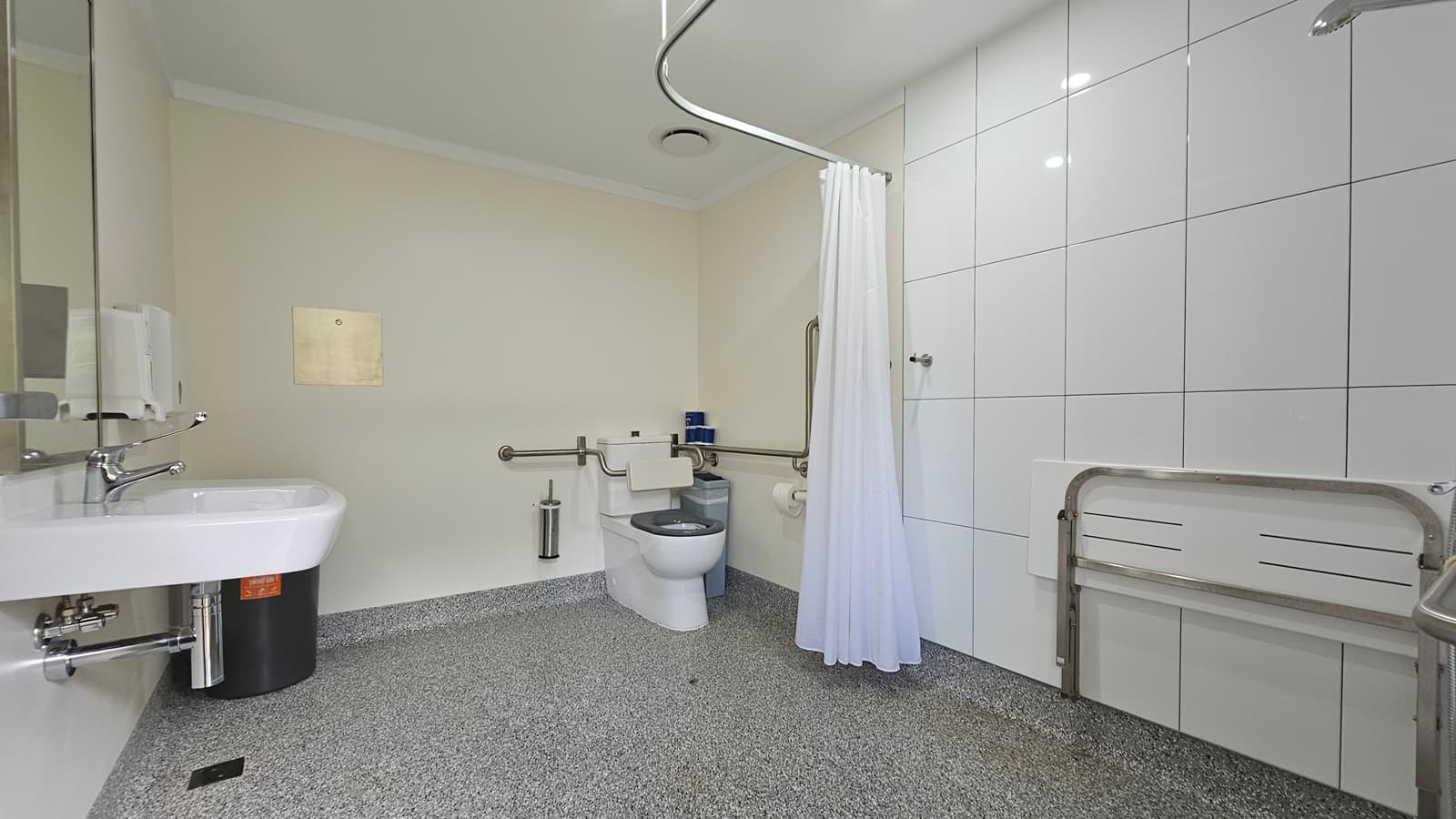 A spacious toilet with a shower. A hand basin, mirror, liquid soap, and tissue dispenser are mounted on the left wall, with a black rubbish bin adjacent. To the right is a toilet bowl equipped with hand grabs, with a toilet brush, rubbish bin, and tissue dispenser nearby. Adjacent is a shower and a foldable bench mounted to the right wall. A white shower curtain hangs from the ceiling. The walls and ceiling are all white, and the floor has grey tiles.