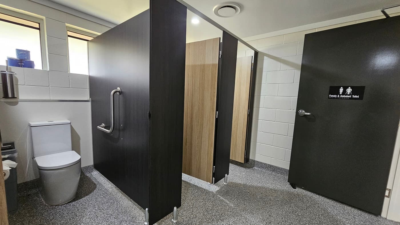 Three bathroom stalls, each with a black wooden door and timber partitions. To the right is a toilet bowl with handrails. To its left is a rubbish bin and tissue dispenser. Above is a toilet bowl brush and tissue rolls placed on a window ledge. The wall has white bricks, the ceiling is white with LED lights, and the floor has grey tiles. The inward-opening black door bears a sign indicating gender and accessibility use.