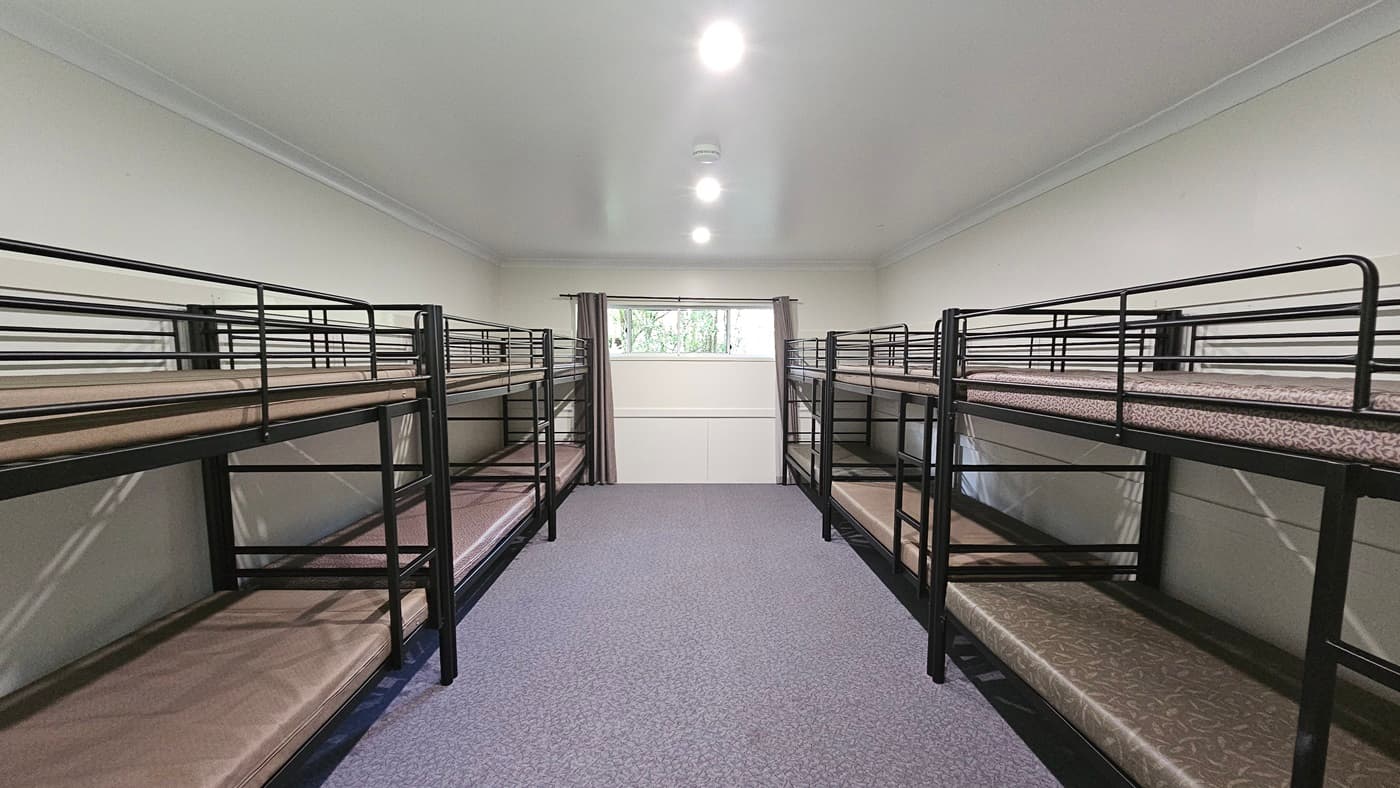 A room with metal bunk beds, each with mattresses, positioned against the walls. In the middle is a glass window with grey curtains pulled to the sides. The walls are white, the ceiling is white with fluorescent lights, and the floor is printed with grey tiles.